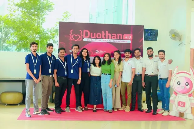 The Duothan 4.0 team lined up on a pink carpet in front of the event backdrop, beside a cut-out of the event mascot