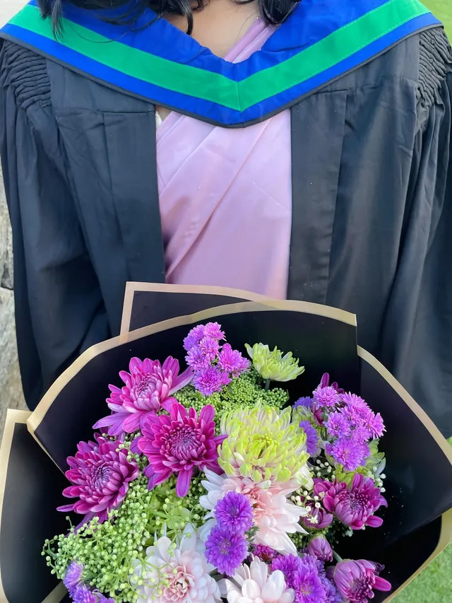 Close-up of a graduation gown with a blue and green hood over a pink saree, holding a bouquet of purple, pink and green chrysanthemums