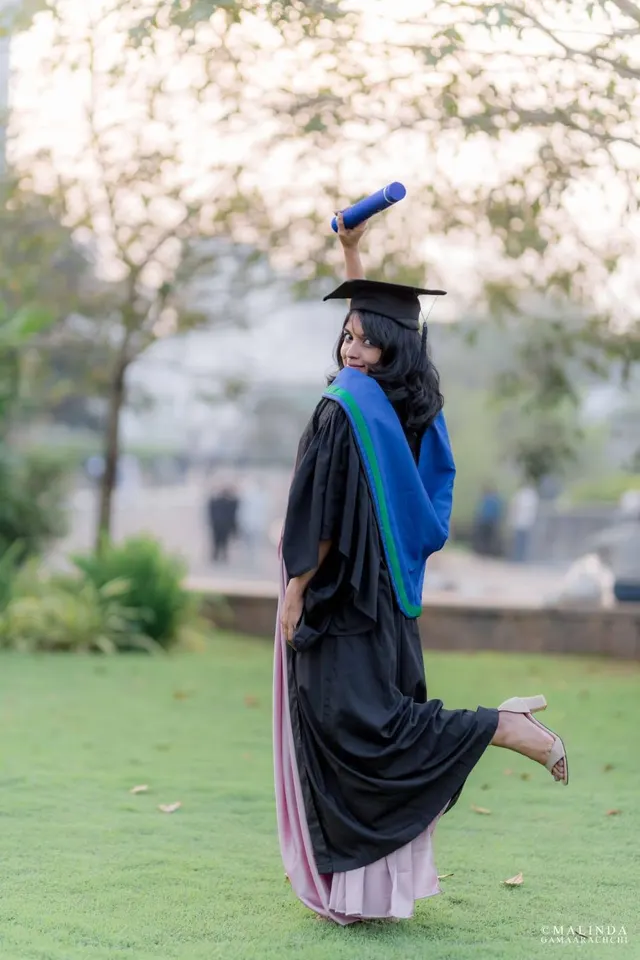 Wathsala in cap and gown on the lawn, looking back over her shoulder with her degree scroll raised and one heel kicked up